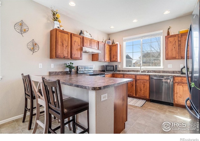 2118 74th Avenue Ct, Greeley, CO 80634 - kitchen featuring a breakfast bar, a peninsula, under cabinet range hood, brown cabinetry, and stainless steel appliances