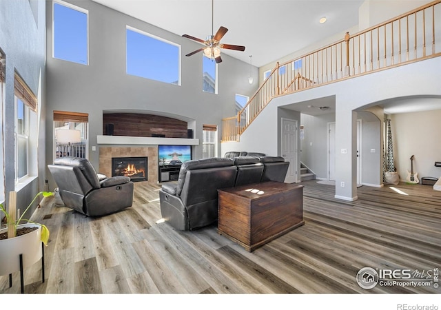 living room with a high ceiling, ceiling fan, arched walkways, a tile fireplace, and wood finished floors