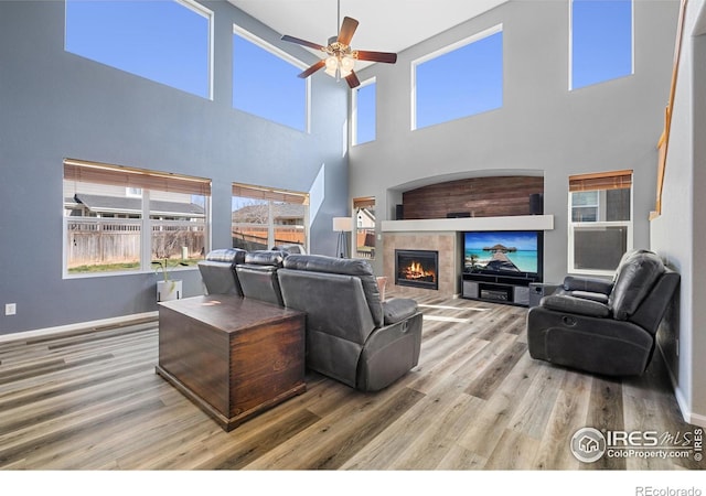 living area featuring baseboards, a ceiling fan, a fireplace, wood finished floors, and a towering ceiling
