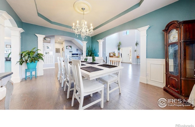 dining area with ornate columns, a raised ceiling, arched walkways, wood finished floors, and wainscoting