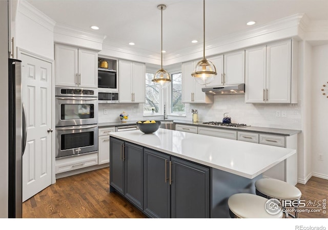 kitchen with appliances with stainless steel finishes, under cabinet range hood, crown molding, dark wood-type flooring, and a kitchen island