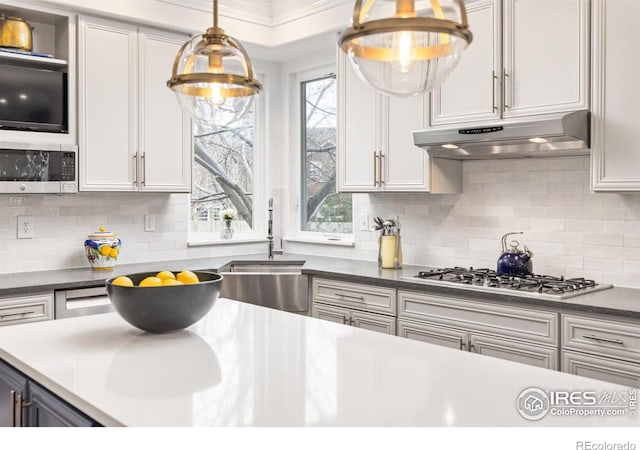 kitchen featuring a sink, under cabinet range hood, stainless steel appliances, pendant lighting, and decorative backsplash