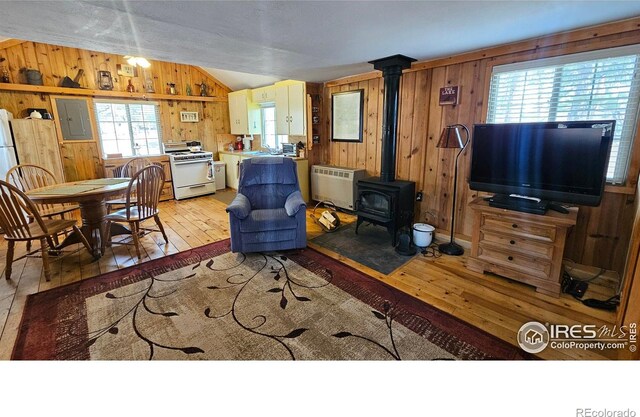 living area with light wood-type flooring, electric panel, wooden walls, a wood stove, and lofted ceiling