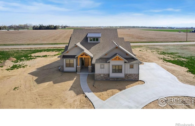 view of front of house featuring a rural view, roof with shingles, stone siding, and concrete driveway