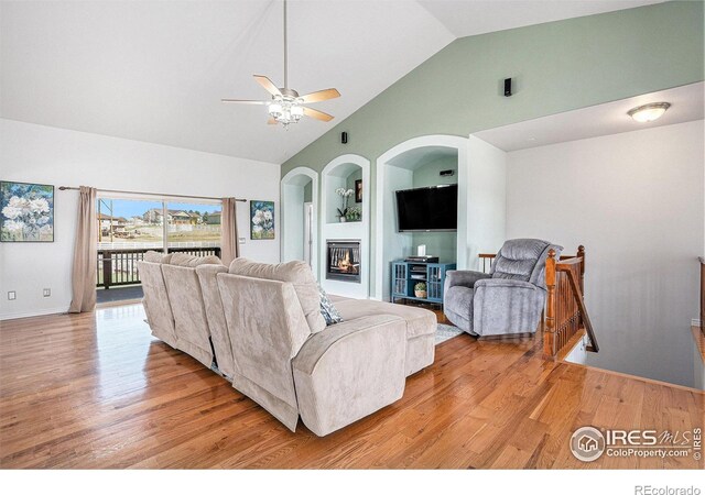living room featuring light wood-type flooring, baseboards, a glass covered fireplace, a ceiling fan, and high vaulted ceiling