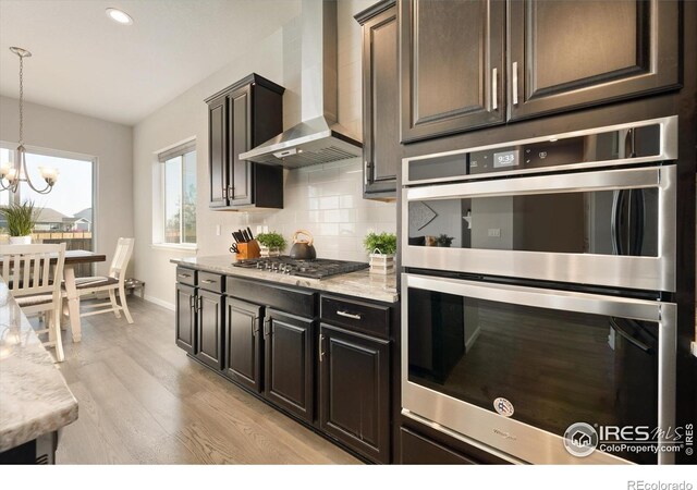 kitchen with appliances with stainless steel finishes, an inviting chandelier, light wood finished floors, wall chimney exhaust hood, and decorative backsplash