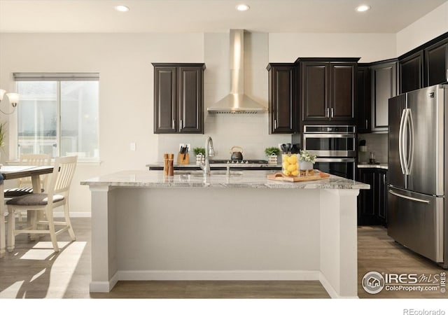 kitchen with appliances with stainless steel finishes, light stone counters, light wood finished floors, wall chimney exhaust hood, and a center island with sink