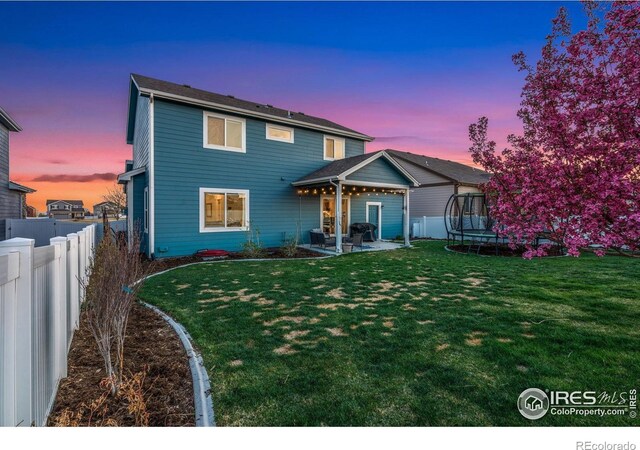 back of house at dusk with a trampoline, a lawn, a patio, and fence