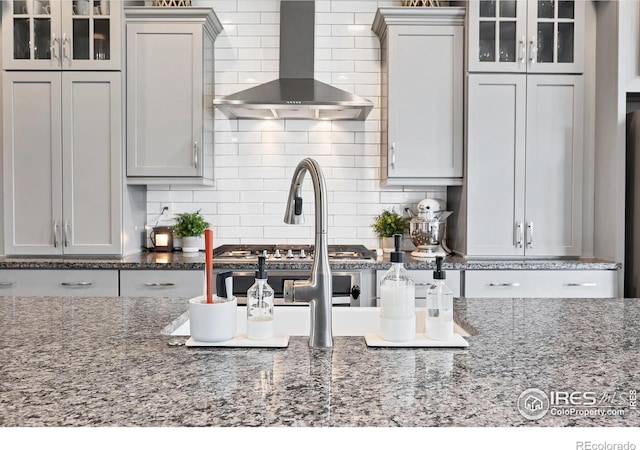 kitchen with glass insert cabinets, backsplash, dark stone counters, and wall chimney range hood