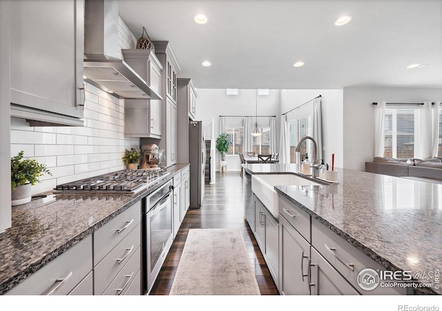kitchen with dark wood-style floors, stainless steel appliances, backsplash, wall chimney range hood, and a sink