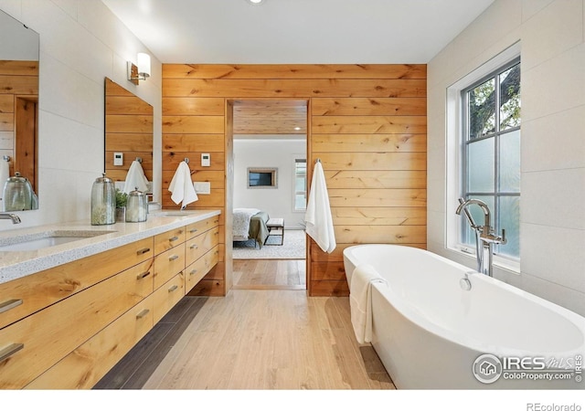 ensuite bathroom featuring double vanity, a freestanding bath, wood finished floors, a sink, and wooden walls