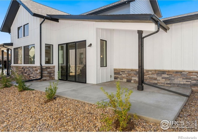 back of property with roof with shingles, stone siding, and a patio