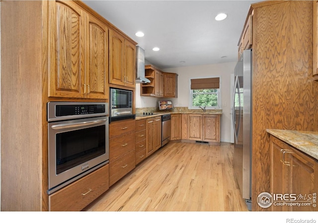 kitchen featuring stainless steel appliances, wall chimney range hood, light wood-style floors, light stone countertops, and brown cabinetry