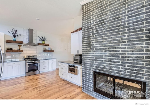 kitchen with wall chimney range hood, appliances with stainless steel finishes, tasteful backsplash, open shelves, and light wood-style flooring
