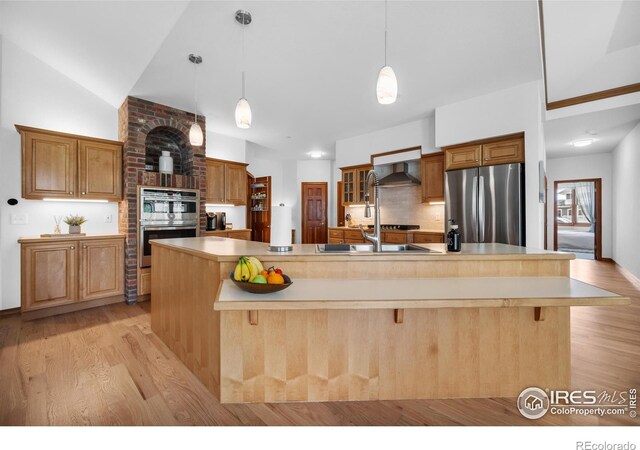 kitchen featuring wall chimney range hood, appliances with stainless steel finishes, light countertops, light wood-style flooring, and decorative backsplash