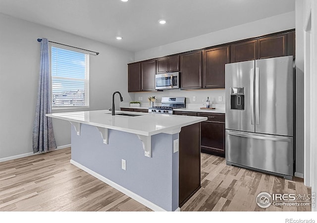 kitchen featuring stainless steel appliances, a sink, light wood-type flooring, dark brown cabinetry, and a center island with sink