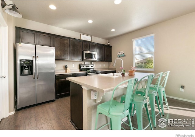 kitchen with wood finished floors, dark brown cabinets, a center island with sink, a sink, and stainless steel appliances