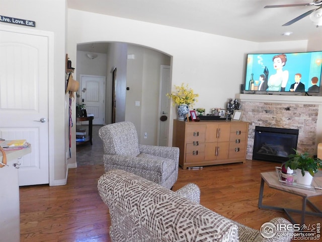 living area featuring arched walkways, a ceiling fan, wood finished floors, and a stone fireplace