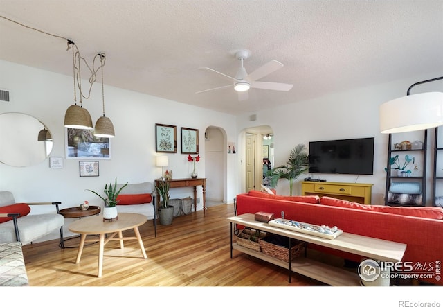 living room featuring a textured ceiling, ceiling fan, visible vents, arched walkways, and light wood-style floors