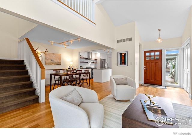 living area with a towering ceiling, visible vents, stairway, track lighting, and light wood-style flooring