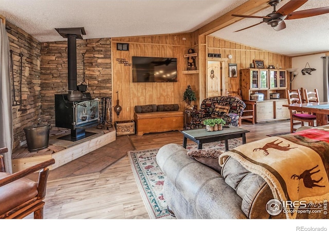 living room with wooden walls, lofted ceiling with beams, a wood stove, a textured ceiling, and a ceiling fan
