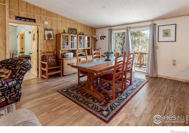dining room with a textured ceiling, light wood-type flooring, baseboards, lofted ceiling, and wooden walls