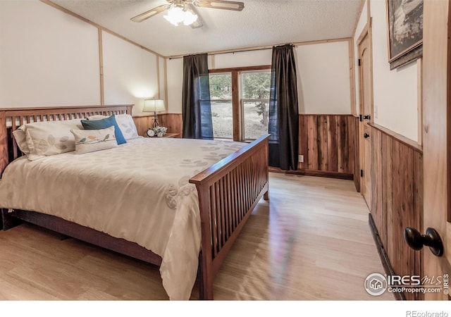 bedroom with ceiling fan, a textured ceiling, wainscoting, wood walls, and light wood-style flooring
