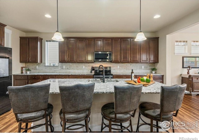 kitchen with backsplash, appliances with stainless steel finishes, a kitchen island with sink, and hanging light fixtures