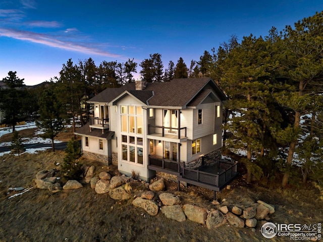 back of property at dusk featuring a shingled roof, a chimney, and a balcony