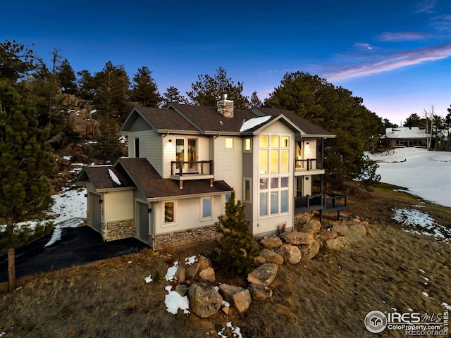 view of front of home featuring a balcony, stone siding, roof with shingles, a chimney, and driveway