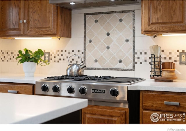 kitchen featuring stainless steel gas stovetop, ventilation hood, decorative backsplash, brown cabinets, and light countertops