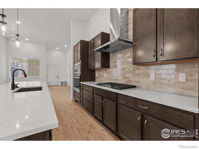 kitchen featuring wall chimney range hood, appliances with stainless steel finishes, light wood-type flooring, dark brown cabinets, and a sink