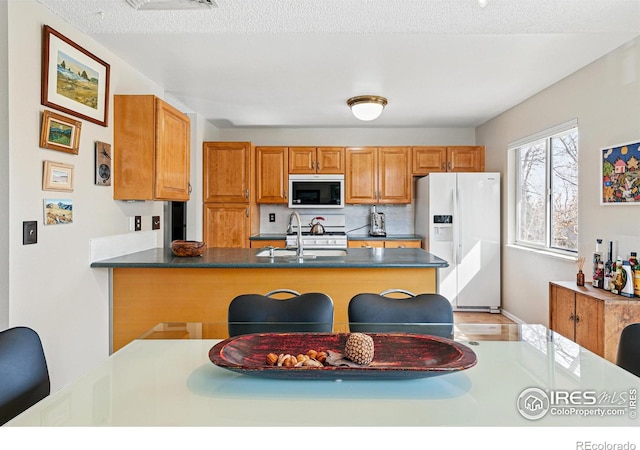 kitchen featuring a peninsula, dark countertops, white appliances, brown cabinets, and a sink