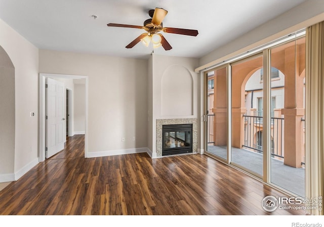 unfurnished living room with a ceiling fan, arched walkways, wood finished floors, baseboards, and a glass covered fireplace