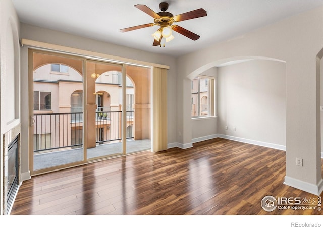 empty room featuring a ceiling fan, arched walkways, wood finished floors, baseboards, and a glass covered fireplace