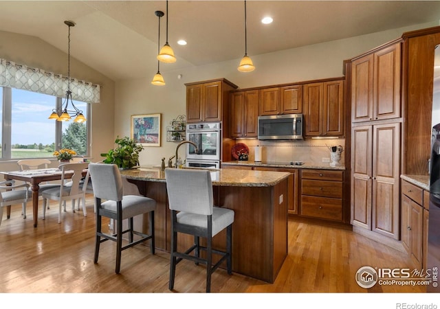 6836 Spanish Bay Dr, Windsor, CO 80550 - kitchen with appliances with stainless steel finishes, a kitchen island with sink, brown cabinetry, stone counters, and lofted ceiling