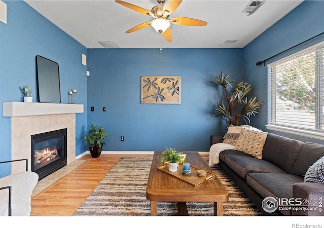 living room with a ceiling fan, visible vents, a tiled fireplace, wood finished floors, and baseboards