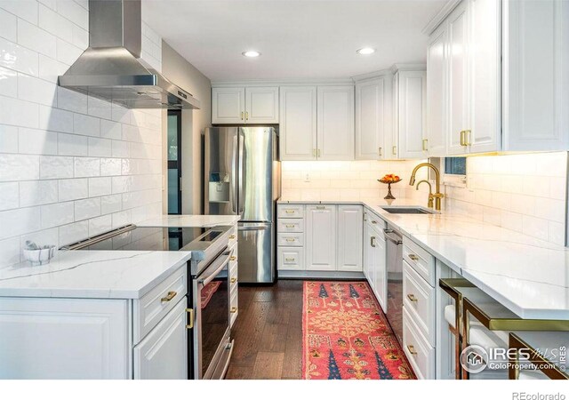 kitchen with wall chimney range hood, appliances with stainless steel finishes, a sink, white cabinetry, and tasteful backsplash