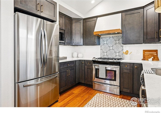 kitchen featuring appliances with stainless steel finishes, vaulted ceiling, light wood-type flooring, light countertops, and premium range hood