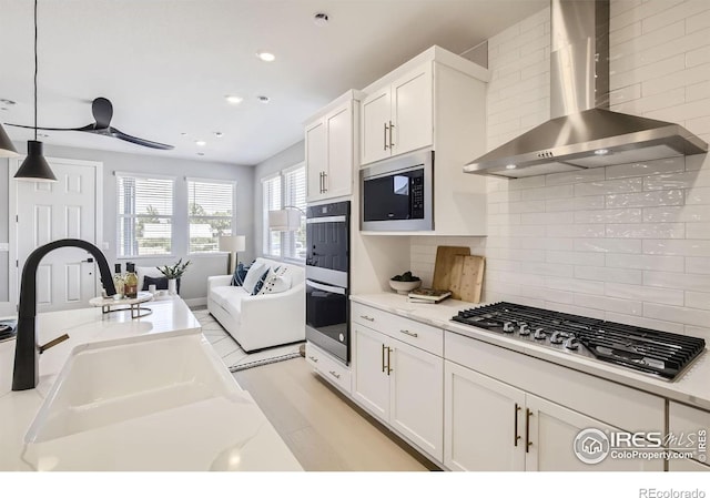 kitchen with wall chimney range hood, hanging light fixtures, a sink, stainless steel appliances, and white cabinetry