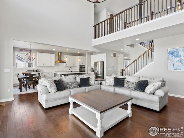 living room featuring dark wood-style flooring, stairway, baseboards, an inviting chandelier, and a high ceiling