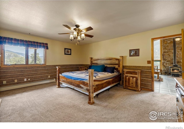 bedroom with light colored carpet, a ceiling fan, a baseboard radiator, and a wood stove