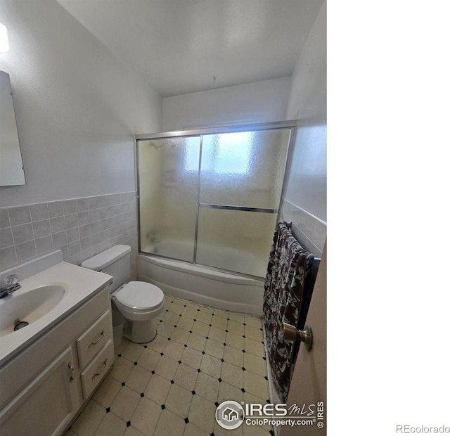 bathroom featuring toilet, tile walls, a wainscoted wall, vanity, and shower / bath combination with glass door