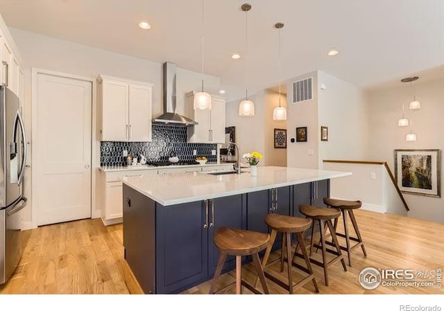 kitchen with a sink, wall chimney exhaust hood, light wood-style flooring, stainless steel fridge, and white cabinets