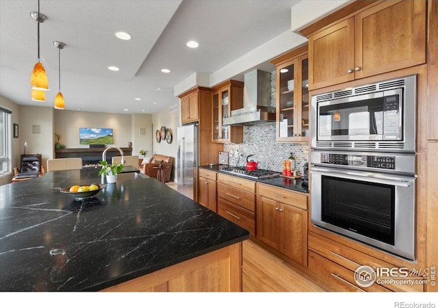 kitchen with glass insert cabinets, brown cabinetry, decorative backsplash, wall chimney exhaust hood, and stainless steel appliances