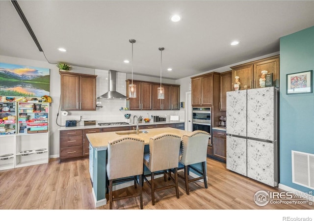 kitchen with wall chimney exhaust hood, appliances with stainless steel finishes, light wood finished floors, an island with sink, and recessed lighting