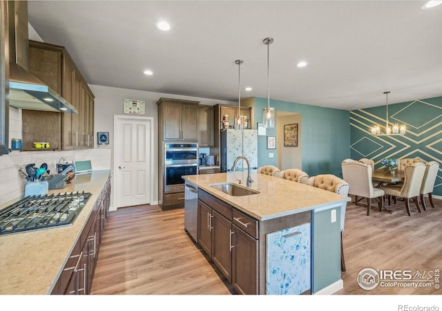 kitchen featuring a sink, a chandelier, stainless steel appliances, light wood-style floors, and wall chimney range hood