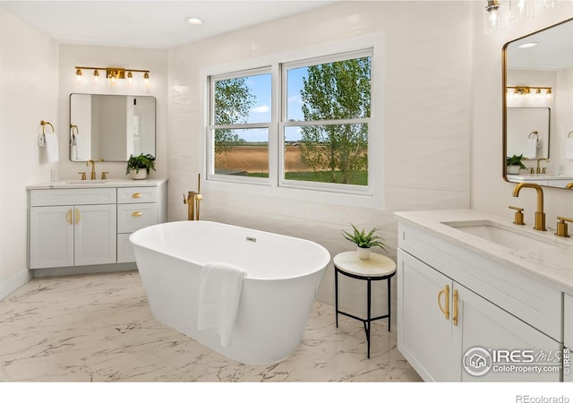 full bath featuring marble look tiles, a freestanding tub, two vanities, and recessed lighting