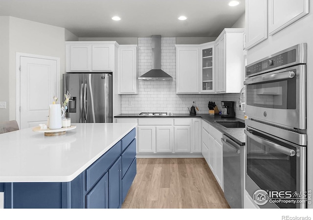 kitchen featuring appliances with stainless steel finishes, blue cabinets, wall chimney range hood, light wood finished floors, and white cabinets