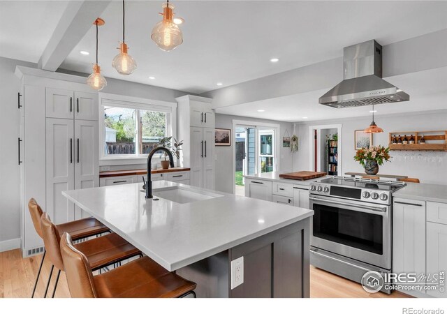 kitchen featuring stainless steel range with electric stovetop, wall chimney exhaust hood, a sink, a center island with sink, and light wood-type flooring